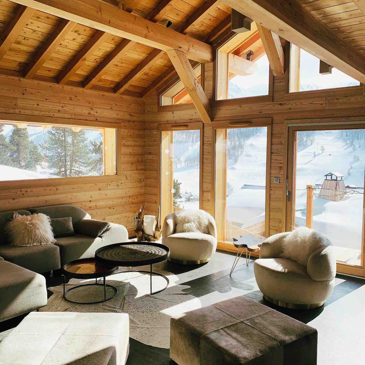A welcoming living area features large windows that flood the space with natural light, showcasing the snow-covered landscape outside. Comfortable seating arrangements include plush sofas and stylish armchairs. A round coffee table sits at the center, complemented by a soft area rug and a simple decorative element.