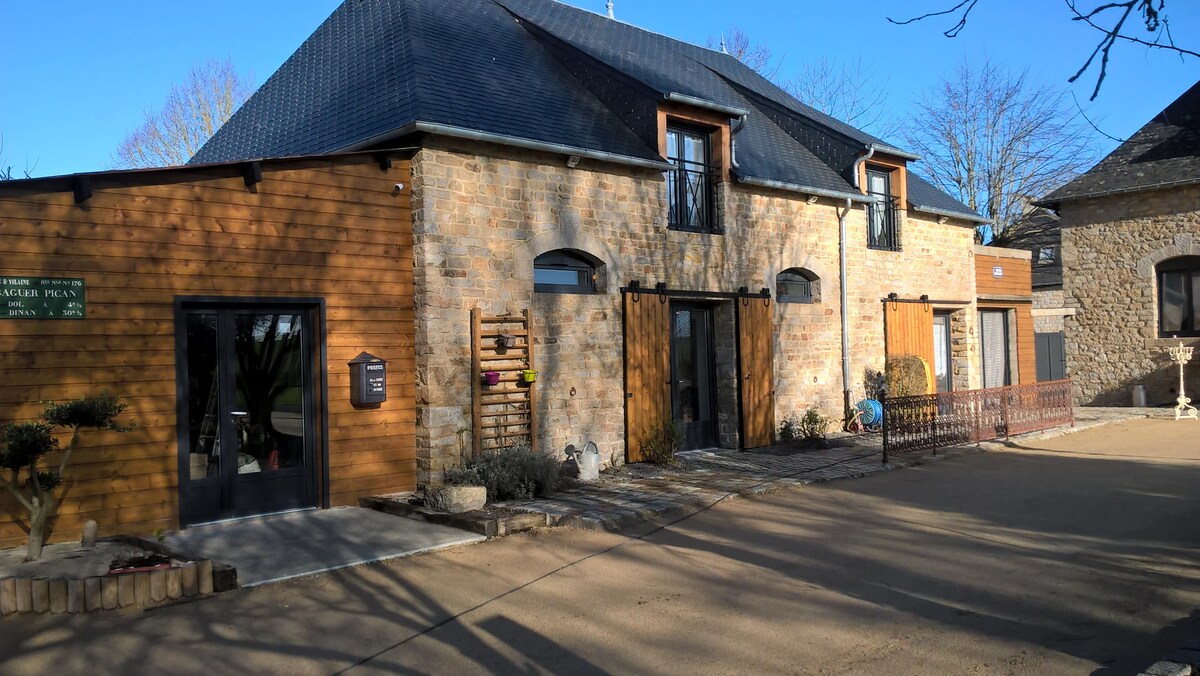 The exterior of the gîte features a mix of brick and wooden siding, with two prominent entrance doors framed by large windows. A paved pathway leads to the entrance, surrounded by landscaped gardens. Clear blue skies above accentuate the tranquil atmosphere of the surroundings.