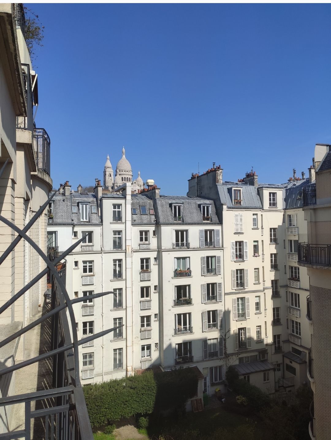 A clear view showcases charming Parisian buildings with slate roofs and balconies. The iconic Sacré Coeur basilica is visible in the distance against a bright blue sky, creating a serene urban scene.