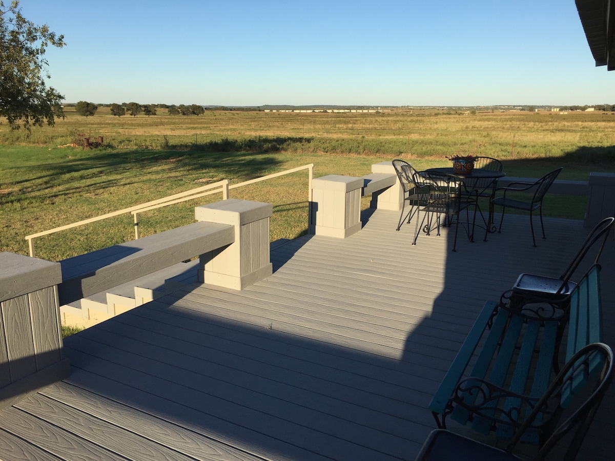 A spacious deck features seating areas with a table and chairs, accompanied by decorative benches. The landscape showcases expansive green fields under a clear blue sky, offering serene views of the surrounding countryside.