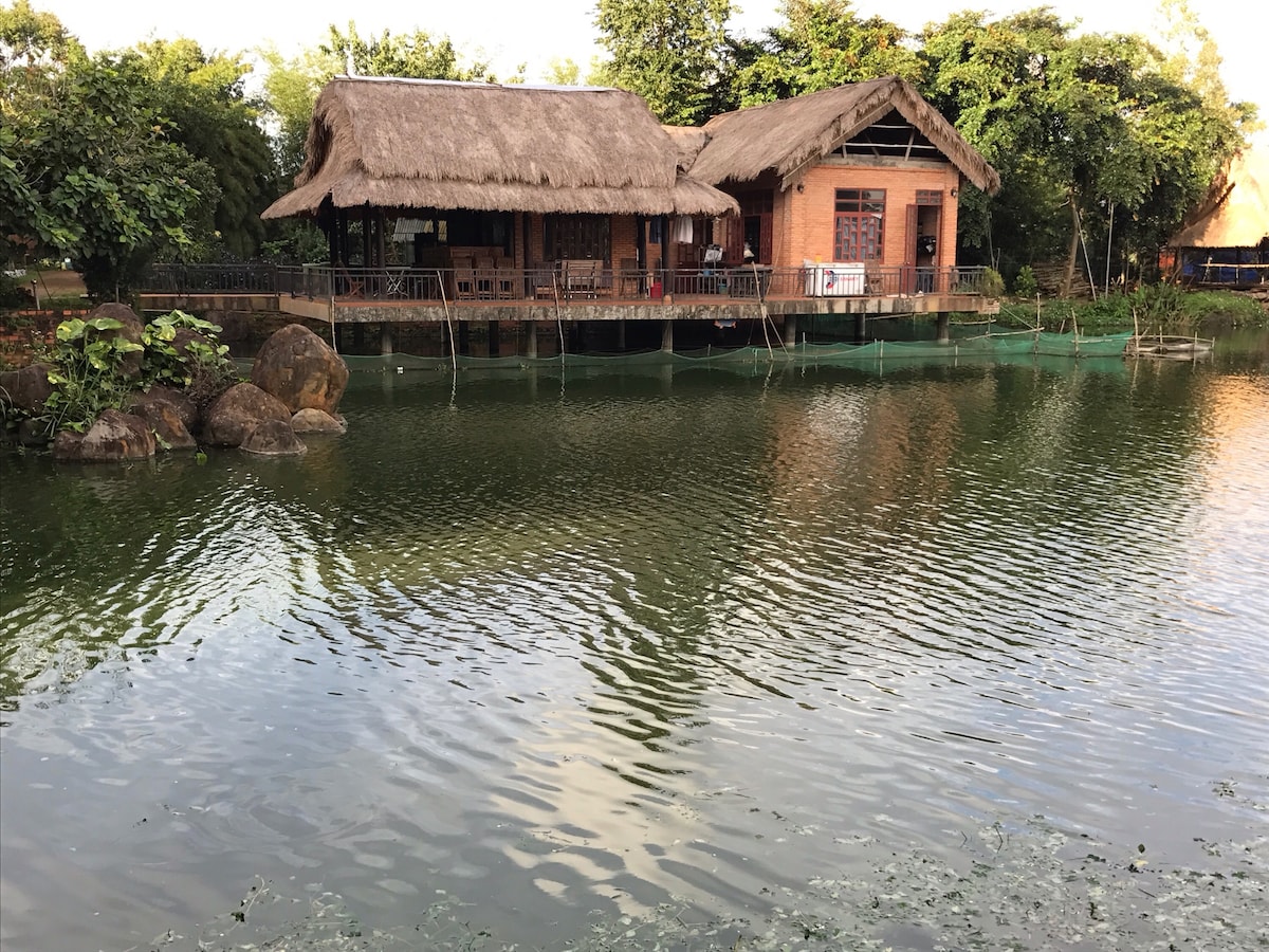 A traditional-style wooden house with a thatched roof sits by a calm pond. Lush greenery surrounds the area, and the tranquil water reflects the building, enhancing the serene atmosphere. A small outdoor deck provides a place to enjoy the natural setting.