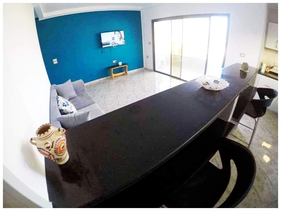 A modern living space is showcased with a blue accent wall and a sleek black countertop. A comfortable seating area features a gray couch, and a television is mounted against the wall. Large glass doors provide access to an outdoor balcony.