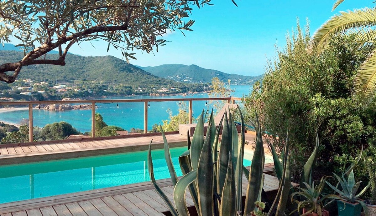 A serene view of the coastline is displayed from a wooden terrace featuring a turquoise pool. Lush Mediterranean vegetation, including agaves and olive trees, frames the scene. The distant mountains complement the tranquil waters, creating a peaceful outdoor atmosphere.