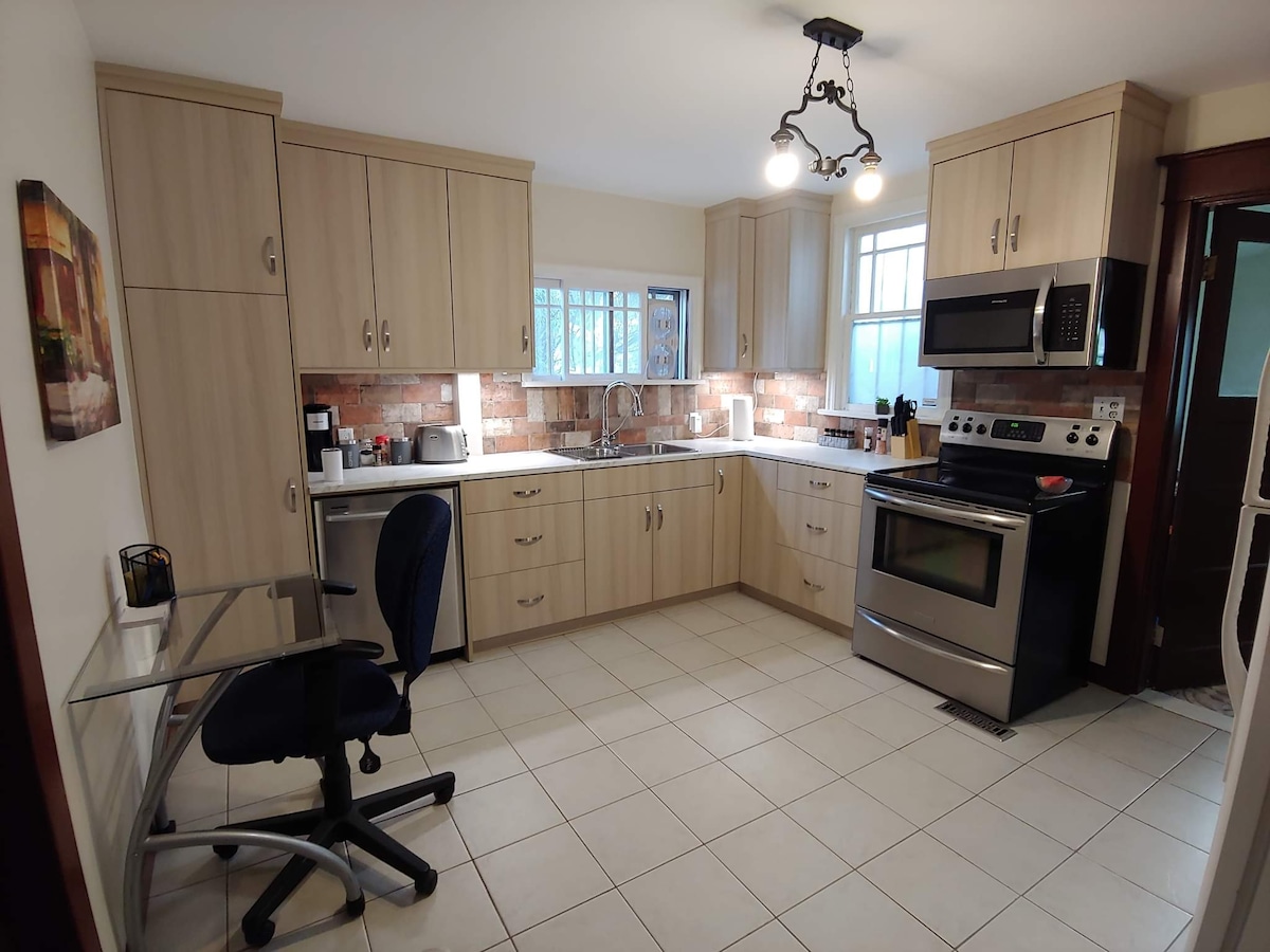 This kitchen features warm-toned cabinetry and stainless steel appliances, including an oven and microwave. A window allows natural light to brighten the space. A small desk is positioned in the corner, and the tiled floor provides a clean, modern appearance.