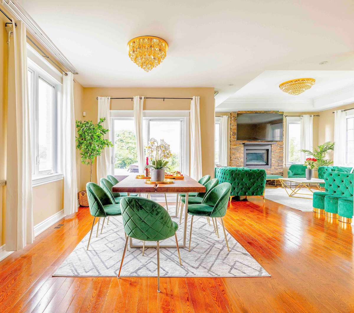 An elegant dining area features a wooden table surrounded by plush green chairs, accented by a chandelier overhead. Sunlight filters through large windows, illuminating the spacious room with warm tones. A stylish seating area with a stone fireplace is visible in the background.