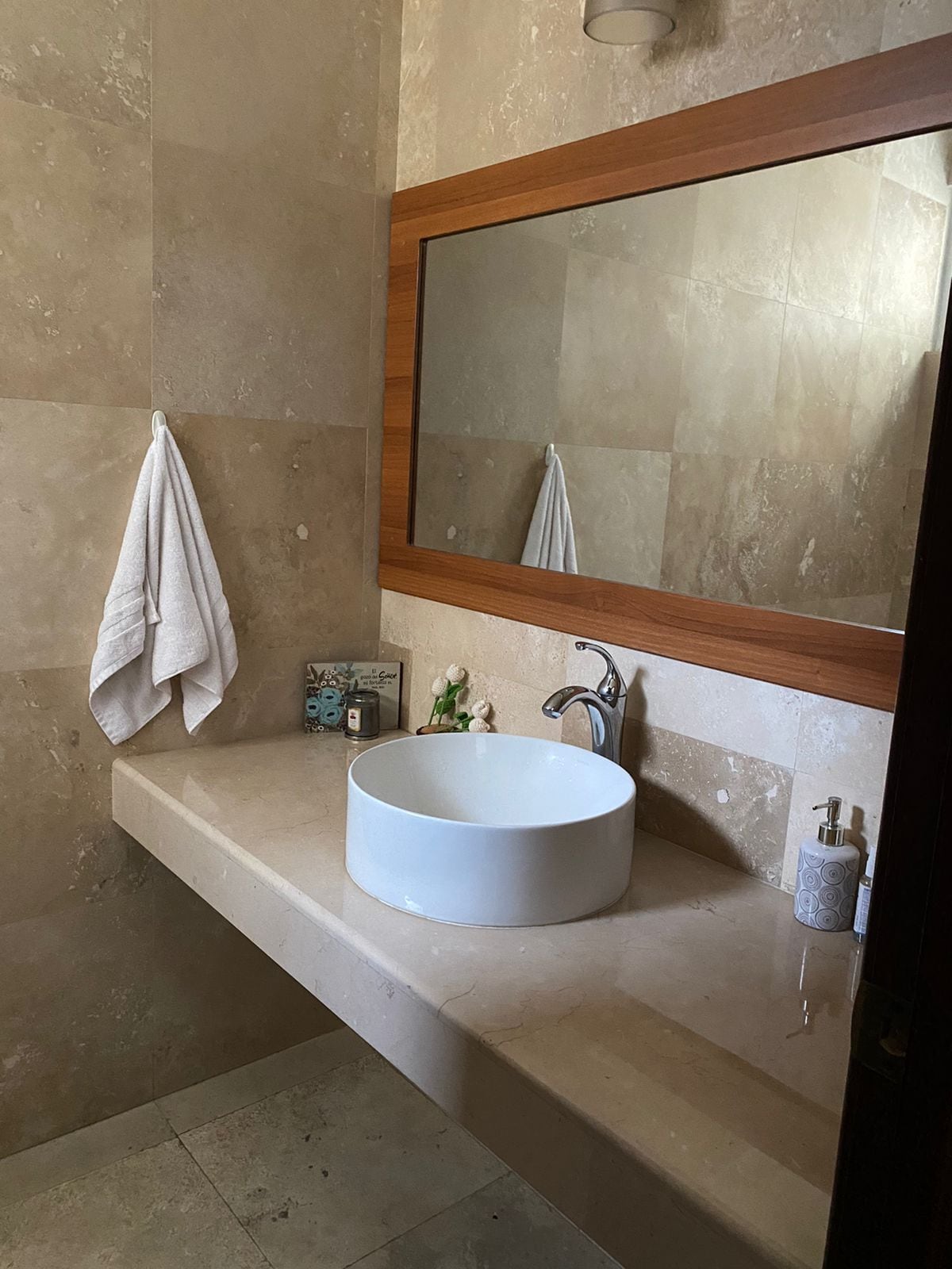 A modern bathroom features a round vessel sink placed on a smooth stone countertop. Natural light reflects off the large mirror above, enhancing the neutral wall tones. A white towel hangs neatly on the side, while decorative items are arranged on the counter.