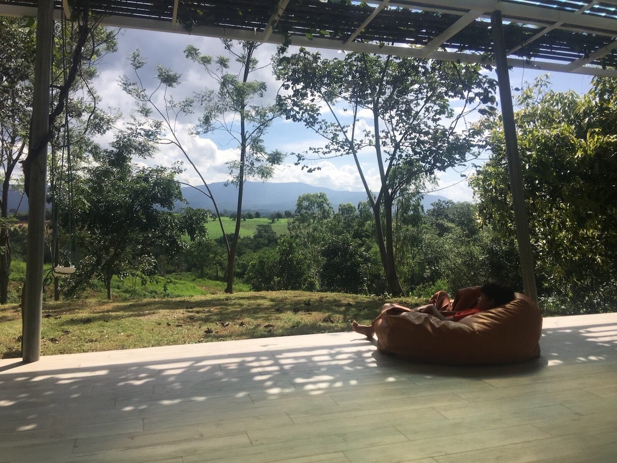 A spacious outdoor area is showcased with a comfortable bean bag chair positioned on a wooden deck. The view reveals a lush landscape of greenery and gentle hills under a cloudy blue sky, creating a serene environment surrounded by nature.