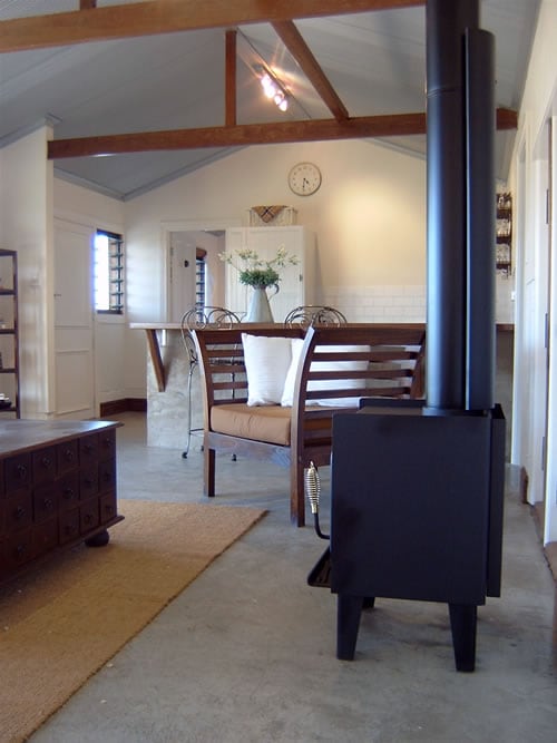 A cozy living area features a black wood stove, providing warmth to the space. Natural light fills the room through large windows, illuminating the wooden dining chairs and a simple table set. Decorative touches include a vase with flowers on the table.