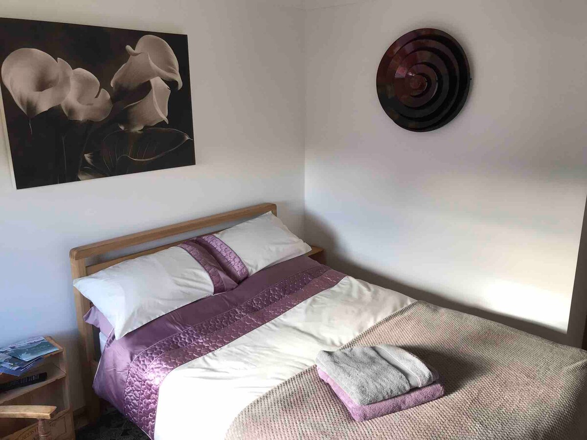 A private room features a wooden bed with a soft white and lavender bedding arrangement. A folded towel is placed neatly on the bed. A piece of wall art depicting flowers is displayed above the bed, complemented by a circular decorative wall piece.