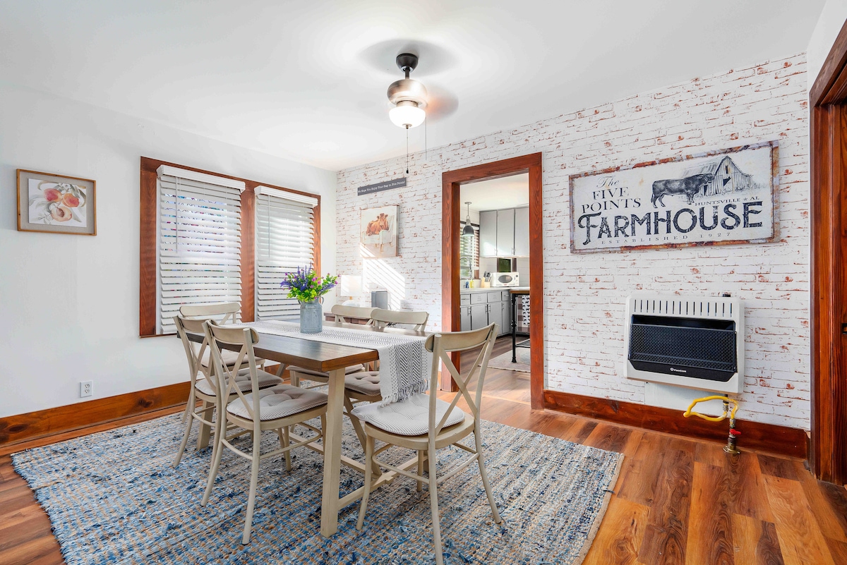A farmhouse-style dining room features a wooden table surrounded by six light-colored chairs. Natural light filters through two windows adorned with plantation blinds. A woven area rug lies beneath the table, and a decorative wall with a vintage-style sign adds character.