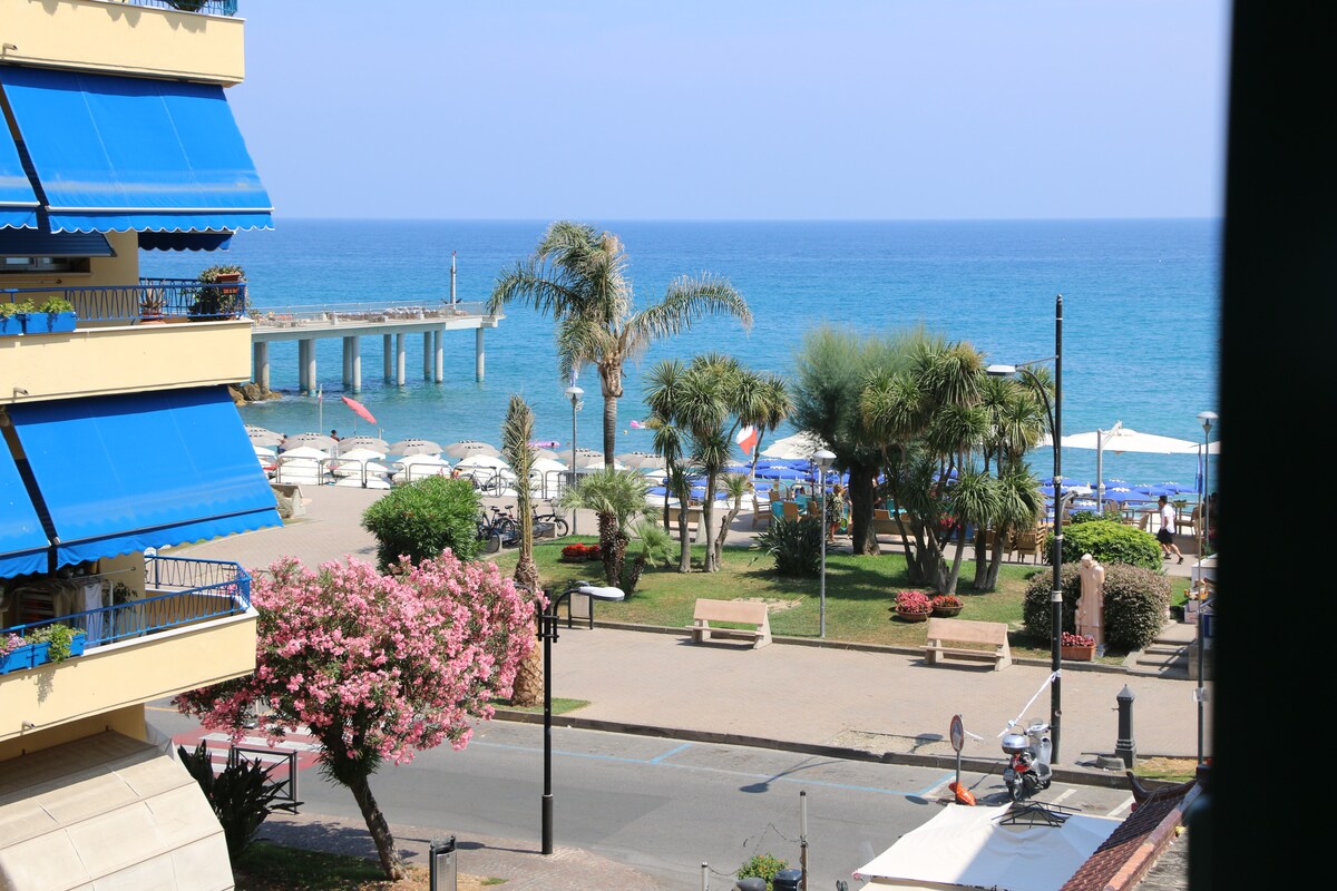 A view of the shoreline reveals a sandy beach with umbrellas and lounge chairs arranged neatly. Palm trees and greenery enhance the scene, while a promenade runs alongside. In the distance, a pier extends into the sparkling water, framed by calm blue skies.