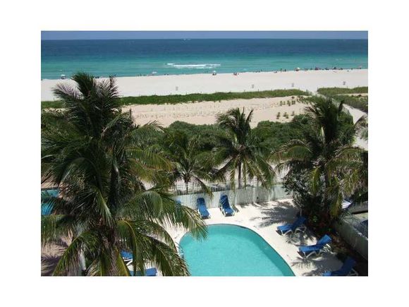A view of a serene pool area surrounded by lush palm trees, leading to the sandy beach. The clear blue ocean stretches into the horizon, where sunlight glistens on the water. Beach loungers are positioned around the pool, offering a relaxed atmosphere.