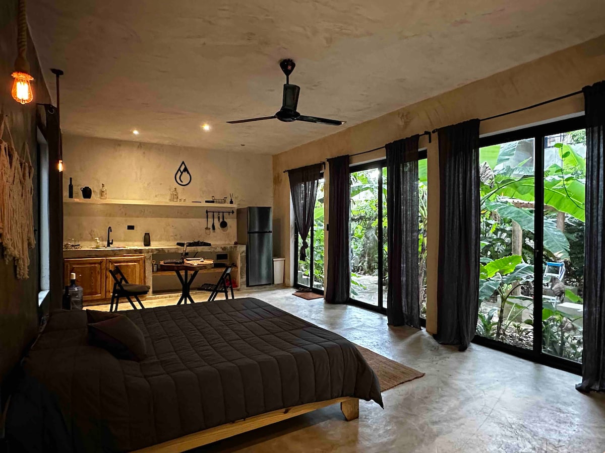 A spacious room features a large bed with a dark blanket. The kitchen area contains wooden cabinetry and includes a dining table for two. Large glass doors allow natural light to illuminate the room, offering views of lush greenery outside.