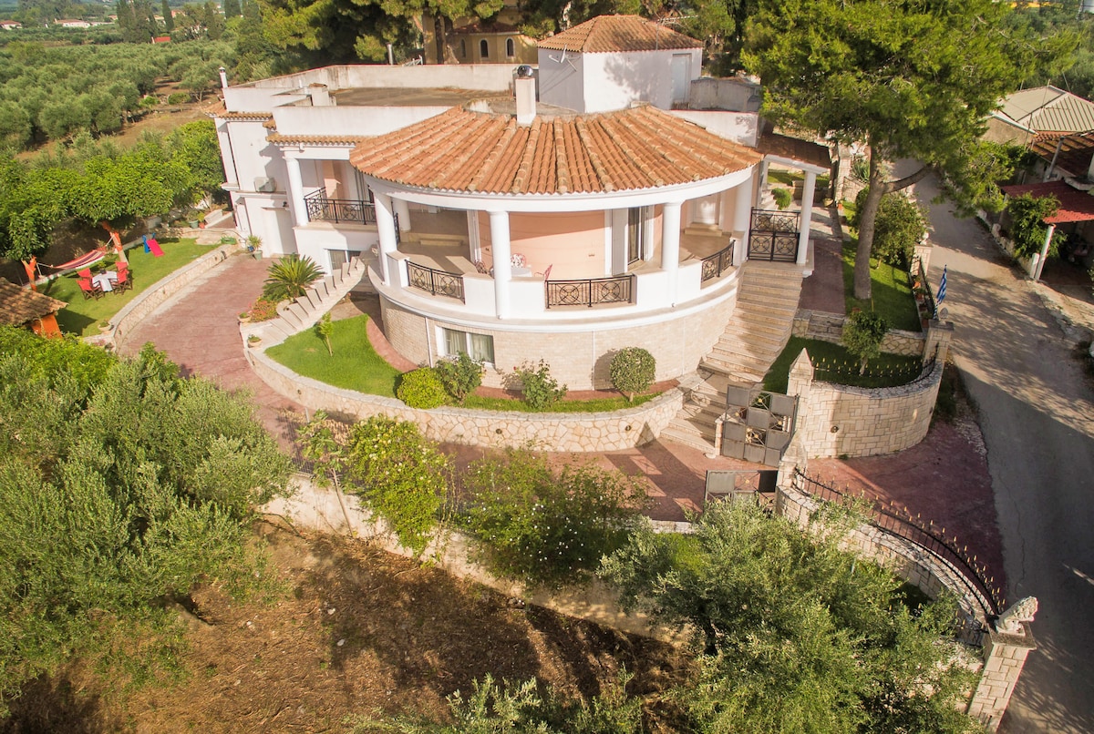 The villa is presented from an elevated perspective, showcasing its unique architecture with a terracotta roof and spacious balconies. Lush greenery surrounds the property, featuring olive trees and well-maintained gardens. A pathway leads to the entrance, emphasizing the serene outdoor space.