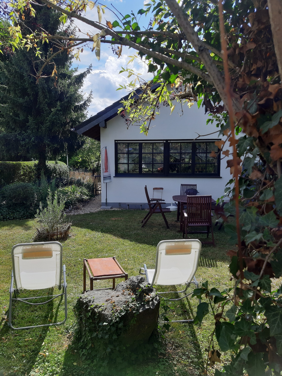A private garden area is depicted, featuring a grassy lawn and various plants. Two lounge chairs are positioned facing a small table, creating an inviting outdoor seating arrangement. The white building with black-framed windows is visible in the background, surrounded by trees.