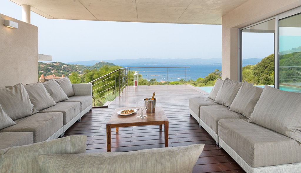 An outdoor lounge area is furnished with expansive, light-colored sofas surrounding a wooden coffee table. The space offers unobstructed views of the Mediterranean Sea, extending to the horizon, and is illuminated by soft natural light.
