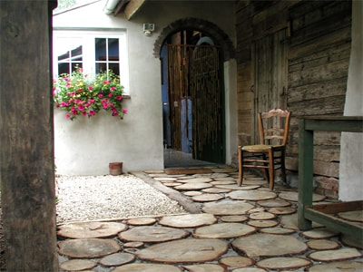 A charming entrance features a stone pathway made of wooden slices leading toward a rustic doorway. Colorful flowers hang from the window, adding a touch of nature. A single wooden chair is positioned nearby, contributing to the welcoming atmosphere.