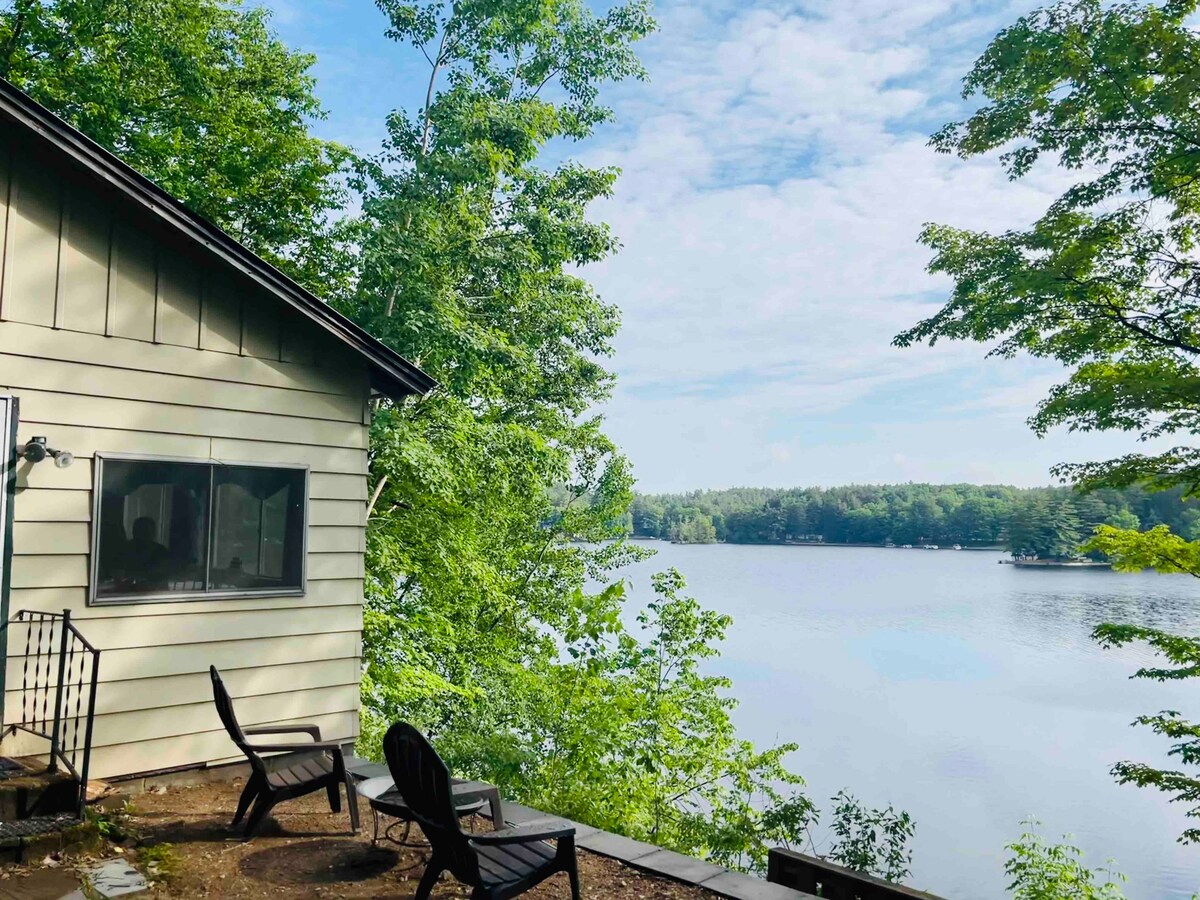 The image captures a serene lake view from the cabin's exterior. Two black Adirondack chairs are positioned on a gravel area, framed by lush green trees. The calm lake reflects the clear sky, offering a peaceful backdrop to the cozy cabin.