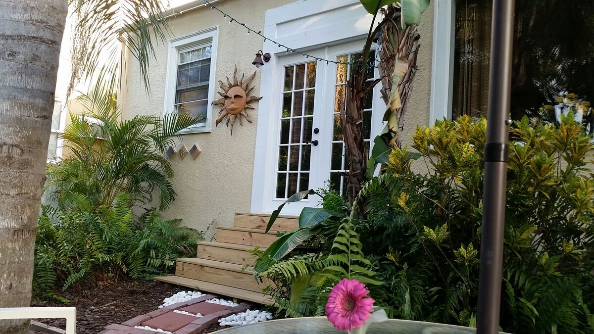 A welcoming entrance features a set of wooden steps leading to bright glass doors. Lush greenery and tropical plants frame the doorway, while a sun motif adorns the wall, providing a cheerful accent to the exterior.