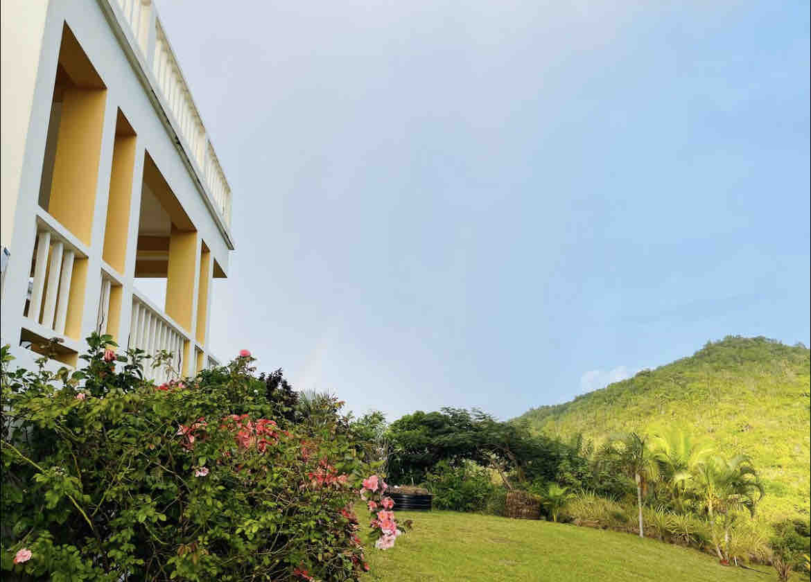 A hillside view features a white structure with a balcony overlooking a landscaped garden. Colorful blooming flowers are in the foreground, while lush greenery and distant hills create a natural backdrop under a clear sky.
