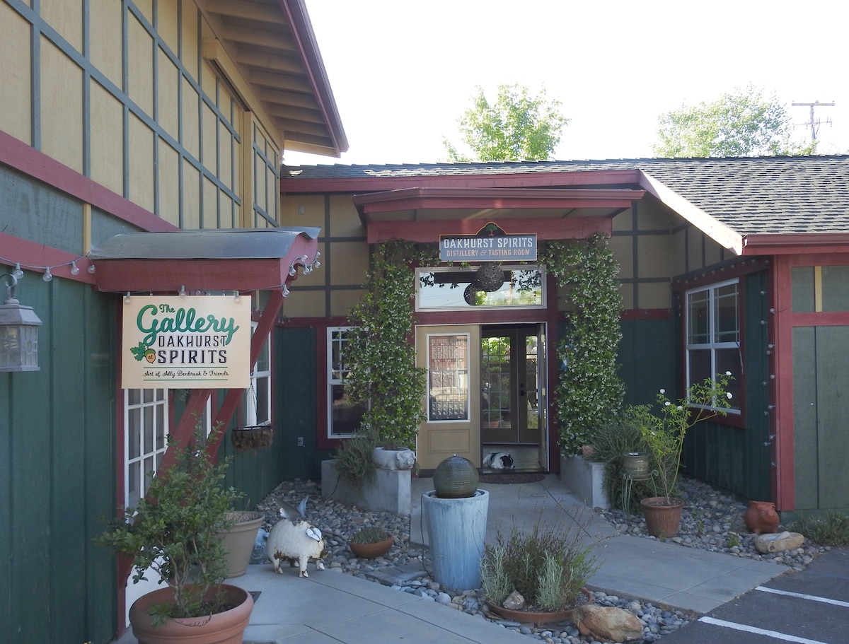 The entrance to Oakhurst Spirits is framed by a vibrant green and red building. Potted plants and decorative stones adorn the pathway, leading to the inviting glass doors. A sign indicating 'The Gallery' hangs prominently, welcoming visitors to the distillery.