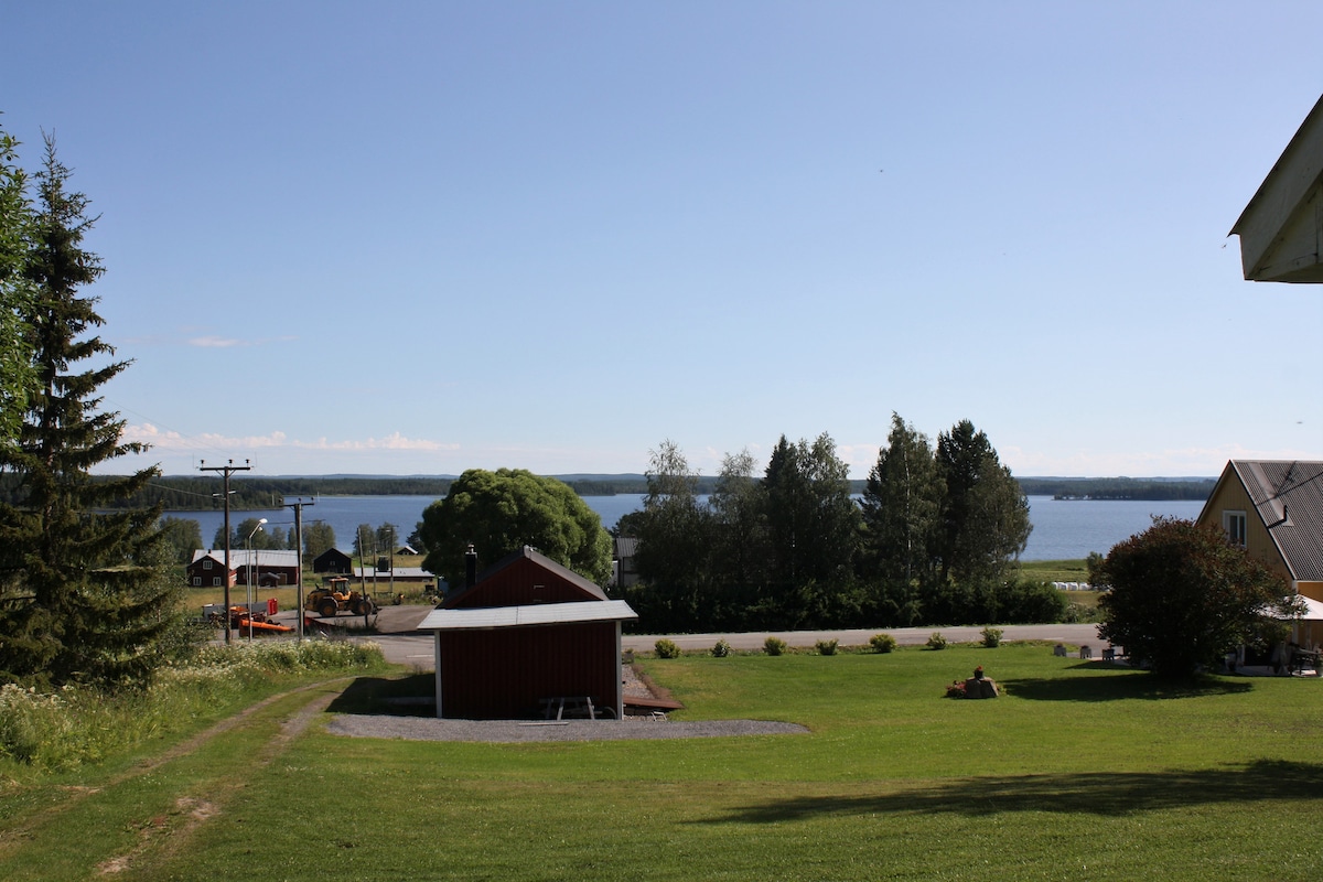The image captures a serene landscape featuring a clear blue sky and Edesjön in the distance. A well-maintained green lawn leads to a cluster of trees, while a red shed is positioned prominently near the water's edge. Nearby structures are visible, enhancing the rural setting.
