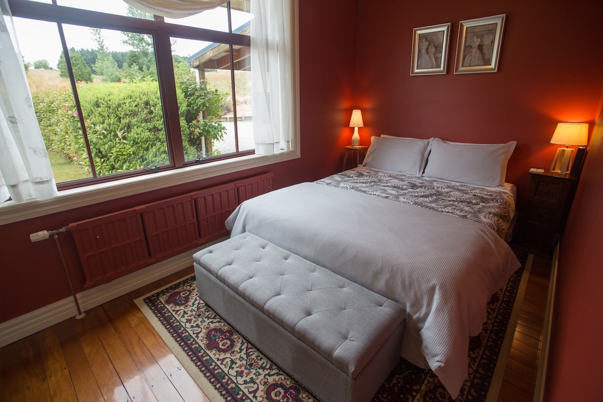 A comfortable queen room features warm red walls and hardwood flooring. A spacious bed with light linens is complemented by bedside lamps. A decorative bench sits at the foot of the bed, and large windows allow for plenty of natural light, overlooking the gardens.