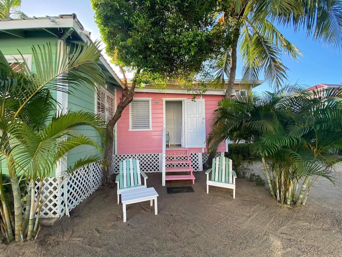 A charming pink wooden cabana is nestled among lush tropical foliage. Two white Adirondack chairs are positioned on sandy ground, inviting relaxation. A small step leads to the front door, framed by vibrant greenery, creating a tranquil outdoor sitting area.