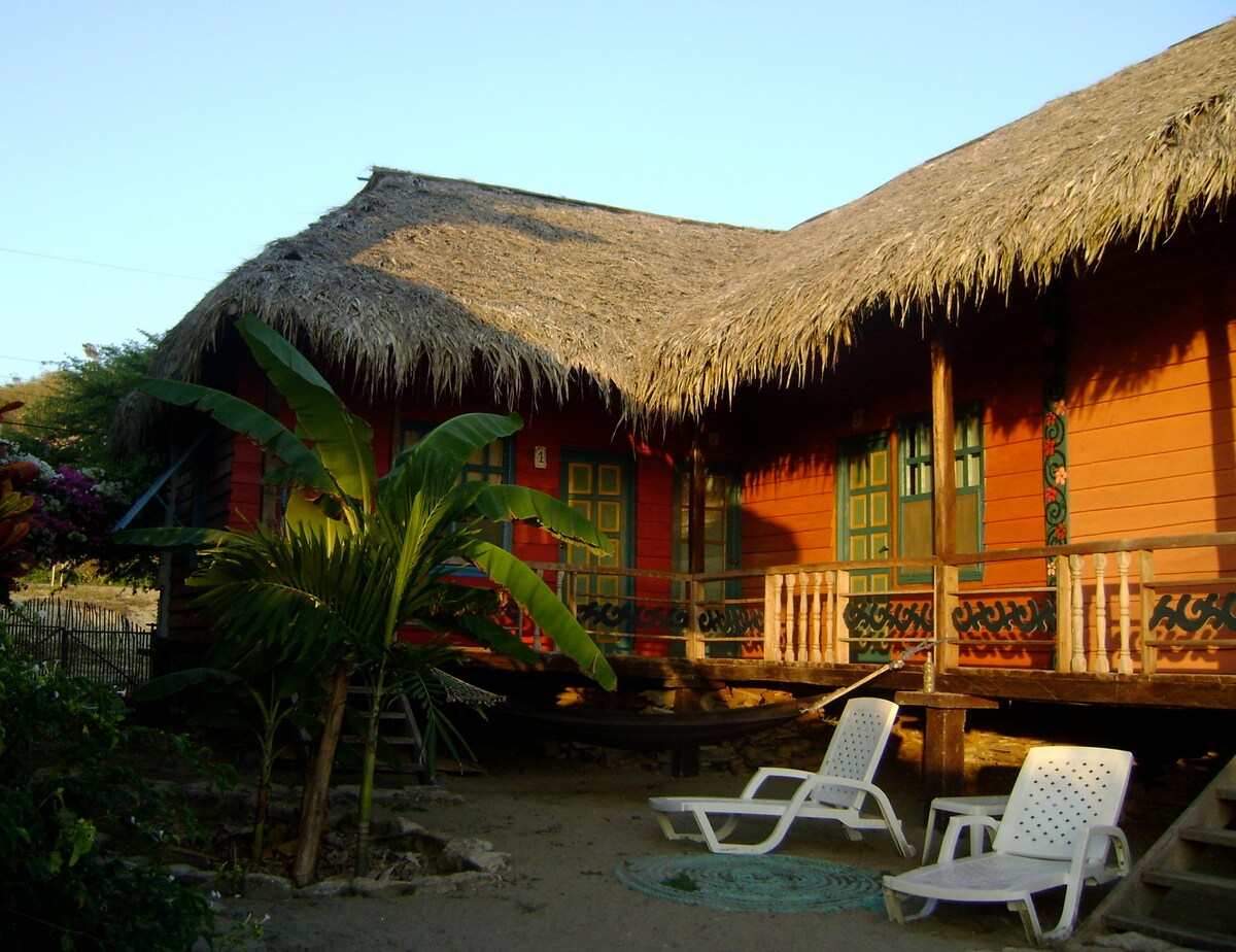 A beachfront wood cabin features a thatched roof and vibrant wooden walls, with a porch adorned by traditional green doors. Two white lounge chairs are positioned in the outdoor space, accompanied by lush green plants and a hammock, creating a relaxed atmosphere in a coastal setting.