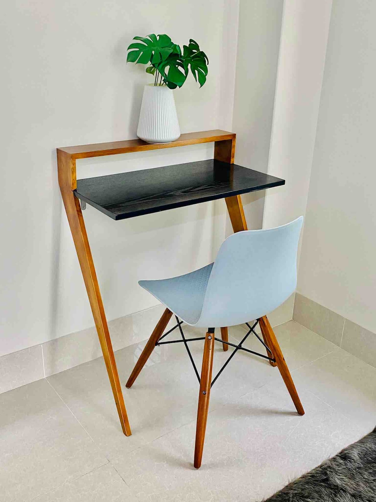 A minimalist workstation is presented with a sleek black tabletop supported by a wooden frame. A light blue chair complements the setup, and a white vase containing a green plant adds a touch of natural decor.