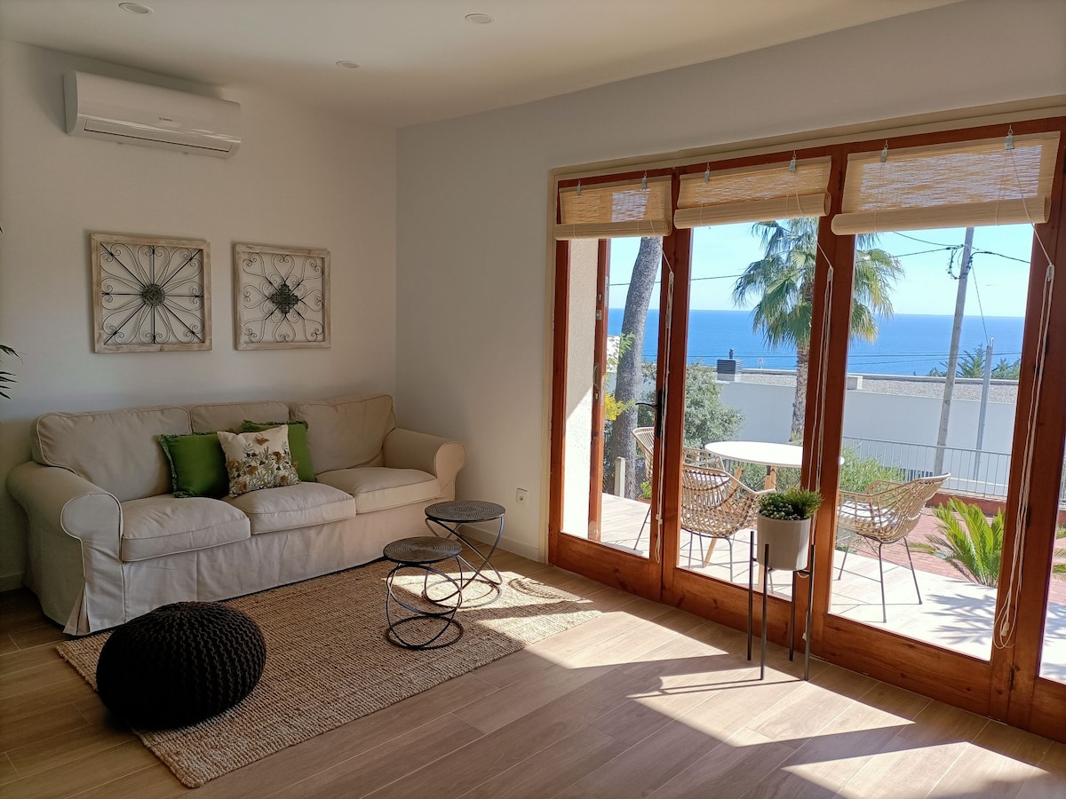 A bright living area features a light-colored sofa adorned with decorative pillows, complemented by a small round table and a low ottoman. Large glass doors provide unobstructed views of the sea and open to a terrace surrounded by palm trees.