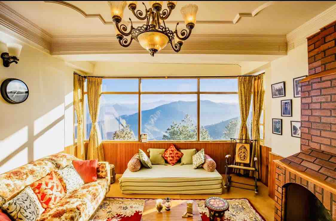 A bright living room is highlighted by large windows offering stunning mountain views. A floral-patterned sofa and a traditional low seat with decorative cushions provide seating. A warm-toned brick fireplace and wall art create a cozy atmosphere, complemented by soft natural light filtering through sheer curtains.