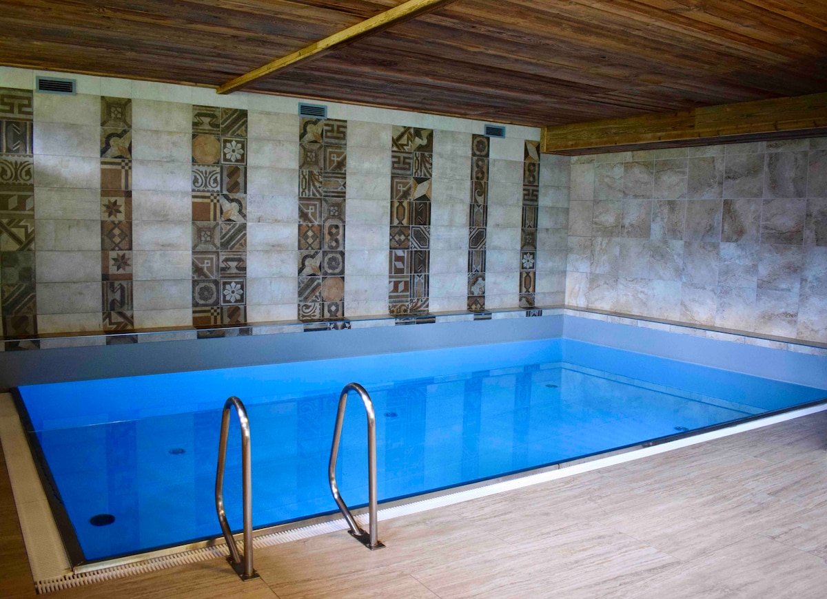 A tranquil indoor pool is featured, with a smooth blue surface reflecting the light. Textured stone decorations line the walls, adding depth to the space. Stainless steel steps lead into the pool, which is surrounded by light-toned tiles and wood accents.