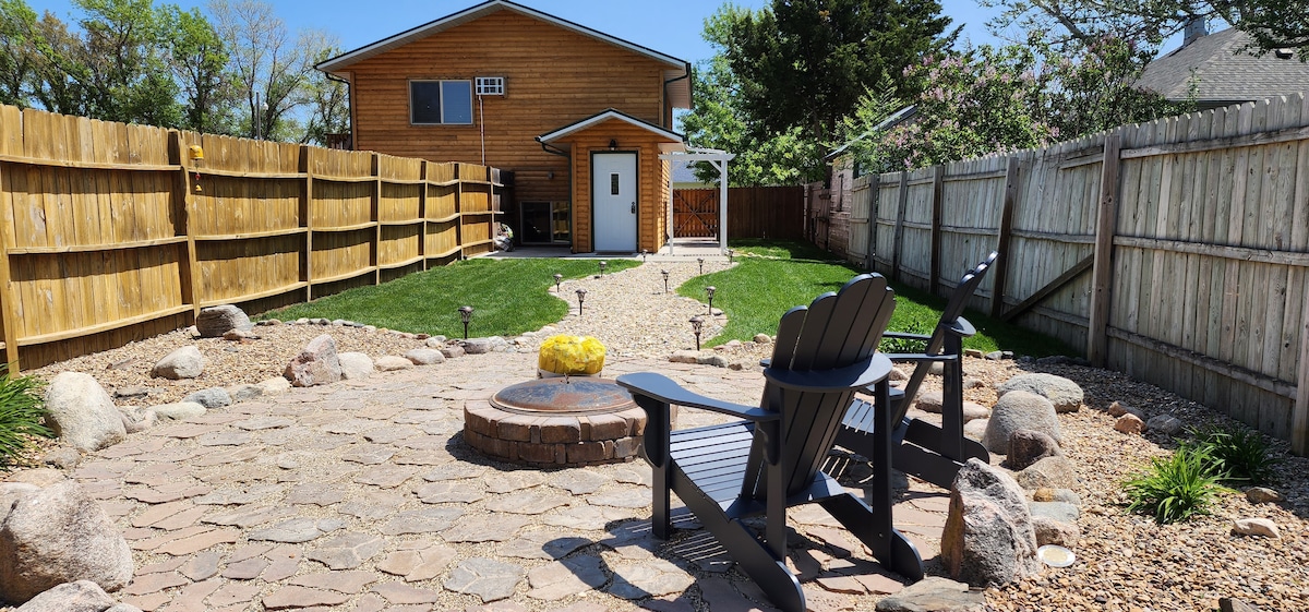 A spacious outdoor area features a circular stone firepit surrounded by wooden chairs. A well-maintained lawn extends into the background, with a patio and a fence providing privacy. The setting is illuminated by decorative lights, creating a cozy atmosphere for evening gatherings.