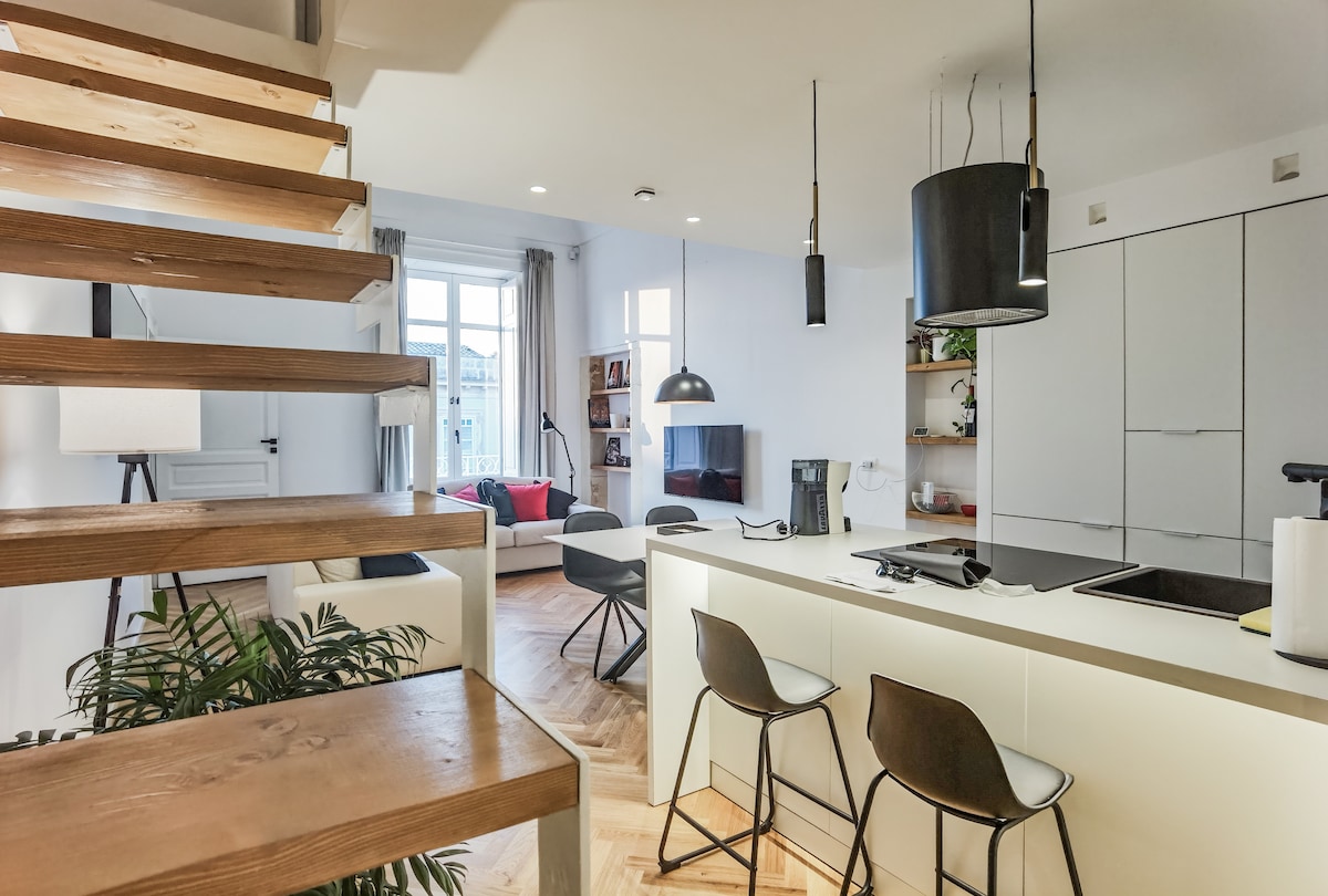 An open-concept space features a modern kitchen island with seating, illuminated softly. Stylish pendant lights hang above, while wooden stairs ascend to additional areas. A cozy living area is visible in the background, showcasing a comfortable sofa and a large window providing natural light.