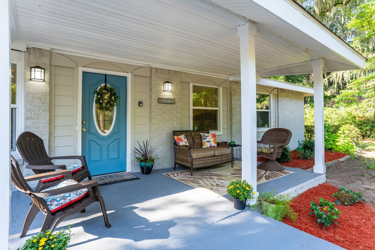 A welcoming front porch features a vibrant blue door and comfortable seating arrangements. Two stylish chairs and a loveseat are arranged on a concrete floor, complemented by potted plants and floral decorations. The surrounding landscaping includes freshly mulched flower beds, creating an inviting outdoor space.