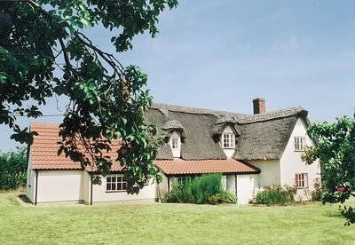 A charming thatched cottage is set amidst lush green surroundings, with a blend of traditional architecture and natural elements. The exterior features a thatched roof, white walls, and wooden window frames. Colorful garden elements can be seen near the entrance, enhancing the inviting presence of the home.