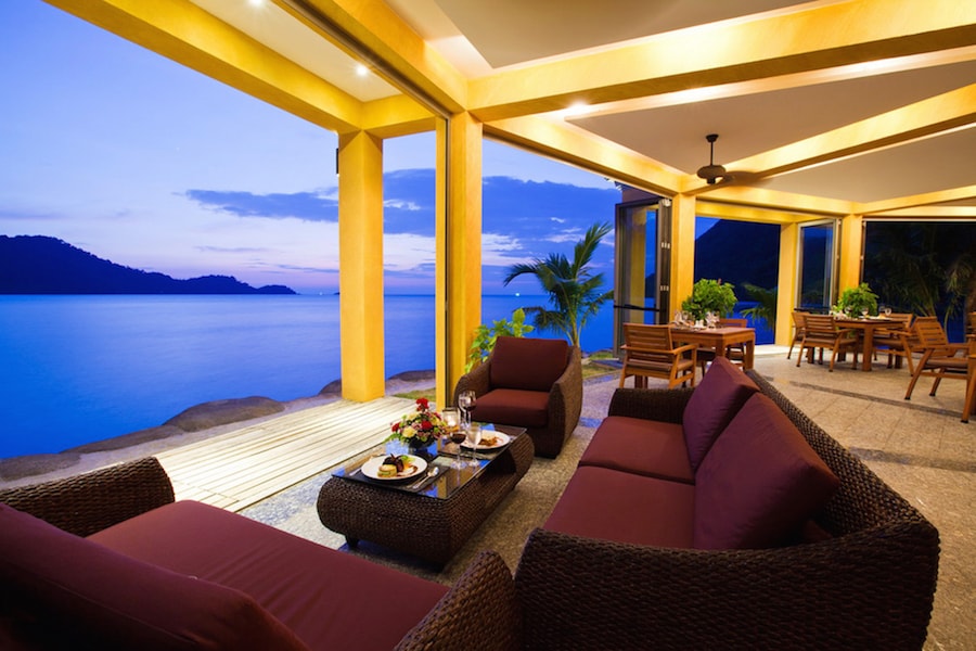 An outdoor seating area is presented with dark wicker sofas and matching armchairs, surrounded by lush greenery. A table is set with food and drinks, offering a view of the calm sea at dusk, with warm colors in the sky reflecting off the water.