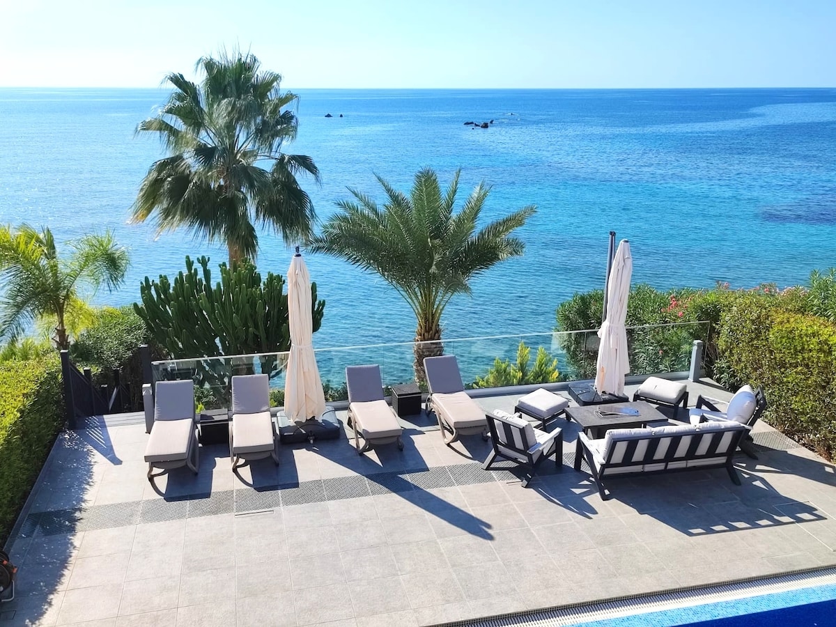An outdoor relaxation area features multiple sun loungers arranged neatly on a tiled deck. Lush greenery surrounds the space, while the calm sea stretches into the distance, providing a serene backdrop. Umbrellas offer shade, and a seating area with a table is positioned nearby.