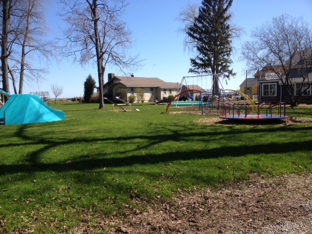 A spacious park area is depicted, featuring vintage playground equipment. A swing set and a merry-go-round are visible on the manicured lawn. Nearby trees provide shade, and several homes are seen in the background, contributing to the welcoming outdoor environment.