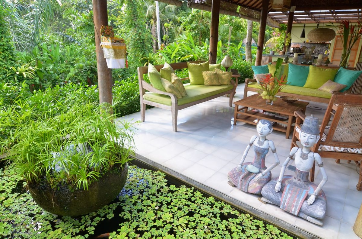 A comfortable seating area features plush cushions in various shades of green and beige, arranged around a low table. Two decorative wooden statues sit beside a tranquil koi pond, adorned with floating lily pads, amidst a lush tropical garden backdrop.