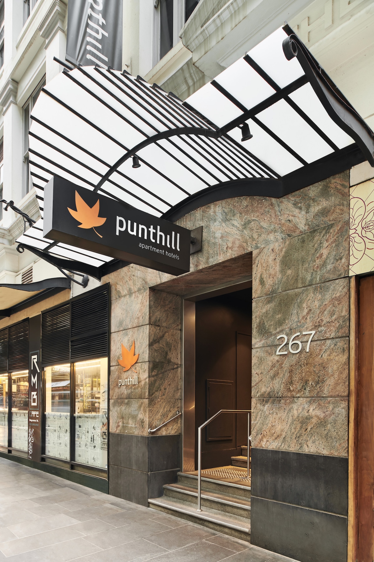The entrance of Punthill Flinders Lane is highlighted by a distinctive awning with a contemporary design. The facade features a combination of marble and stone elements, while the address '267' is displayed prominently beside the entrance. The overall appearance exudes a modern and welcoming atmosphere.