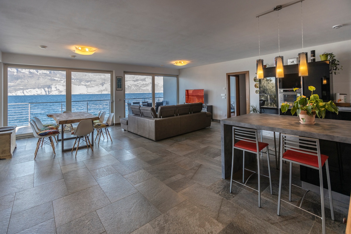 An open space living area features large glass windows overlooking the lake, allowing abundant natural light. A spacious dining table is surrounded by wooden chairs, while a modern kitchen with bar seating is visible. A comfortable seating arrangement offers a relaxed atmosphere, enhancing leisure time.