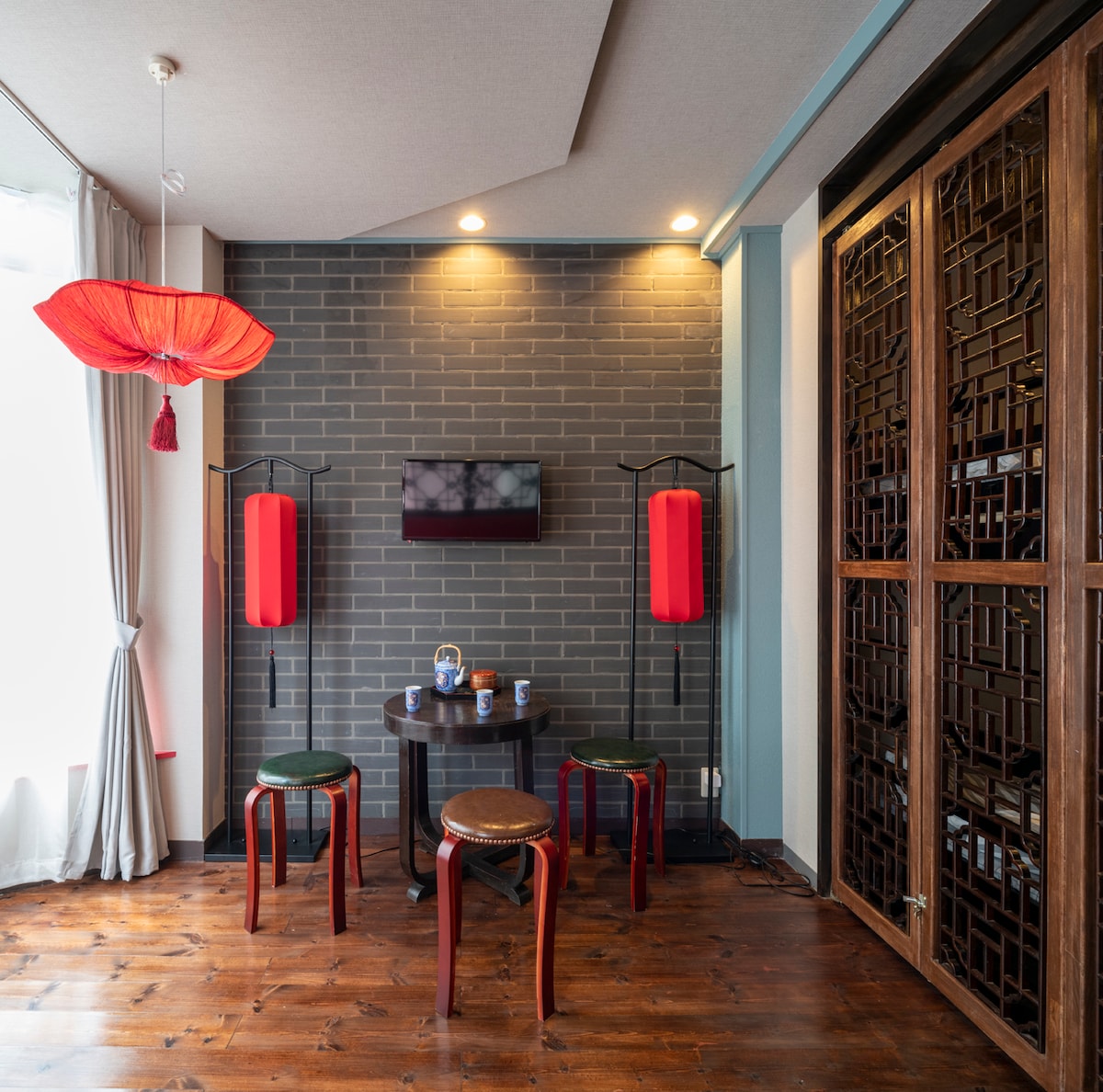 A modern Shanghai-style room features a wooden floor and a gray brick accent wall. Two red lanterns hang overhead, complemented by a black television mounted on the wall. A small dining area is set up with a round table and two red chairs near a decorative wooden screen.