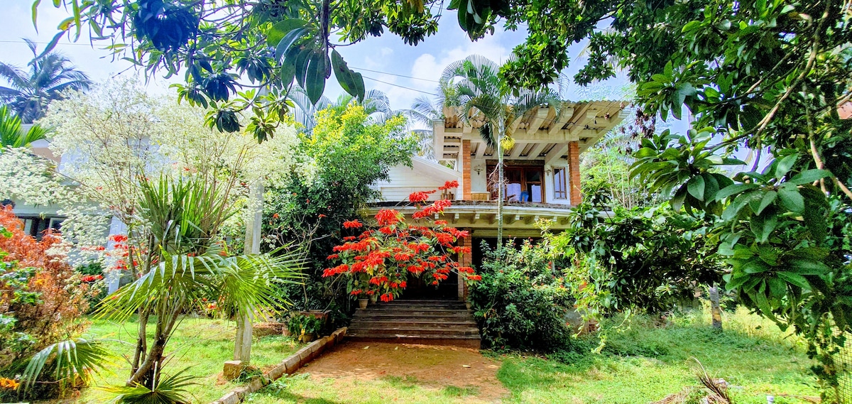 A bungalow is set among lush greenery, featuring vibrant flowered plants in the foreground. The building showcases a porch area with steps leading up, framed by palm and tropical trees, creating an inviting entrance surrounded by nature.