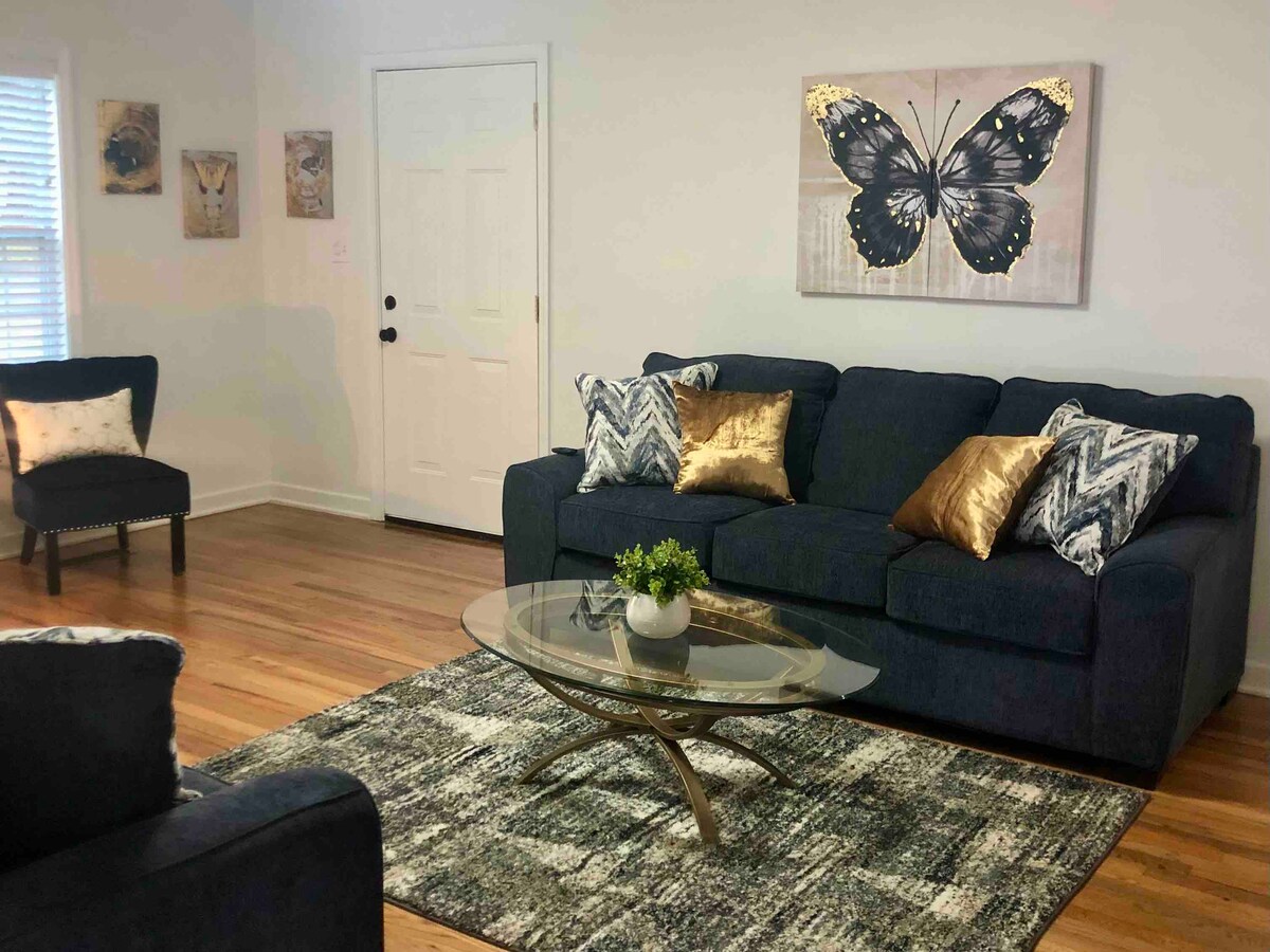 A living area features a dark sofa adorned with textured throw pillows, complemented by an accent chair. A glass coffee table is placed on a patterned rug, with wall art depicting a butterfly enhancing the room's decor. Natural light filters through the window.