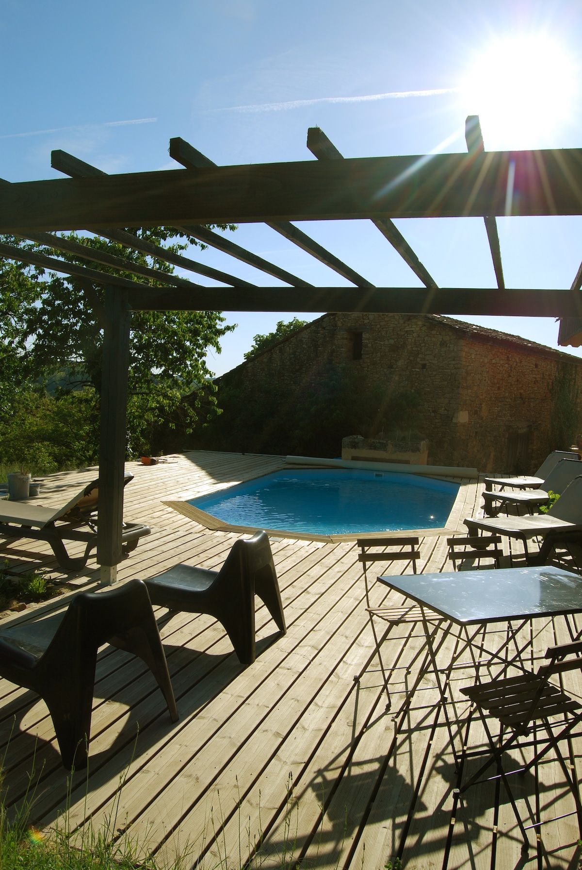 A wooden terrace features a private pool, surrounded by outdoor seating. Sunlight filters through the pergola, casting dappled shadows over the area. A stone structure is visible in the background, enhancing the serene outdoor ambiance.
