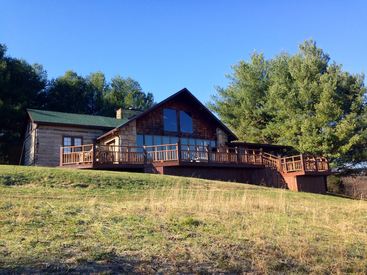 The cabin's exterior is nestled among trees, showcasing a spacious wooden deck with multiple seating areas. Large windows dominate the front, allowing ample natural light into the interior. The surrounding grassy area slopes gently towards the cabin, creating a serene and private setting.