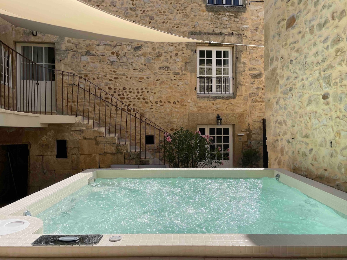 A mosaic spa, measuring 2.30 x 2.30 m, is situated in a private courtyard with stone walls. Sun protection sails are visible overhead, and lush greenery borders the spa, contributing to a serene environment.