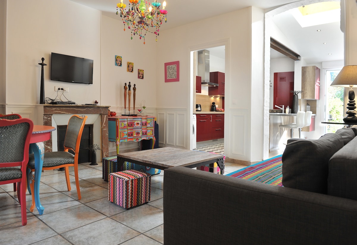 A spacious living area features a dark sofa and a colorful rug. A central coffee table is surrounded by patterned stools. A chandelier with multicolored elements hangs overhead. The area is complemented by a large wall-mounted television and an open view to a modern kitchen.